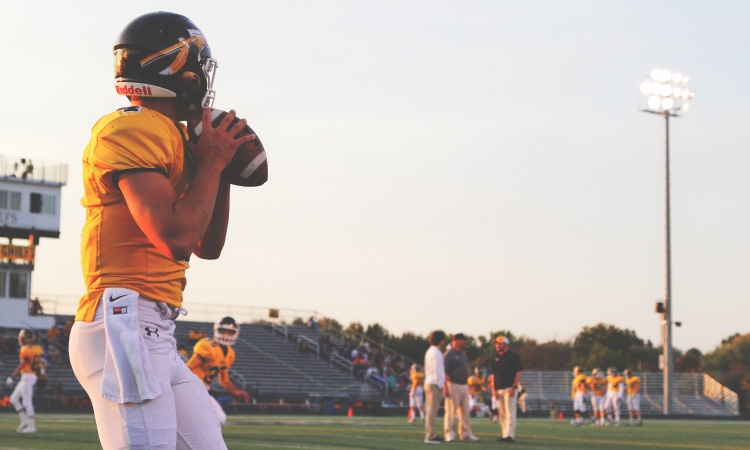 A young man in a yellow football jersey and gear gets ready to throw a football across the field during practice