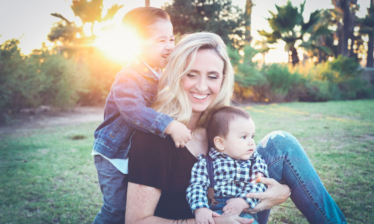 Blonde middle-age woman sits with her baby boy on her lap and her toddler son embracing her from behind as the sun sets