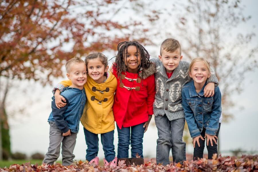 A group of children wearing cold-weather coats smiling outside together by fallen leaves
