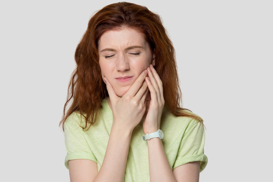 Red-haired young woman in a pale green shirt cringes in pain and touches her cheeks due to sensitive teeth